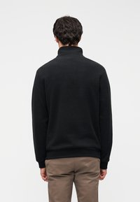 Μαύρο, απαλό φλις πουλόβερ με ψηλό γιακά. Διαθέτει μακριά μανίκια και ριμπ τελείωμα. Εμφανίζεται από πίσω, φορεμένο πάνω από μπεζ παντελόνι.