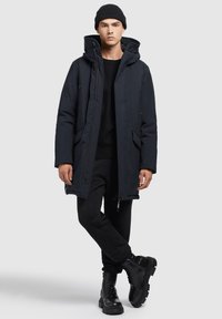 Jachetă parka navy cu glugă, închidere cu fermoar și două buzunare frontale. Pulover negru și pantaloni slim asortați cu cizme negre cu șireturi.