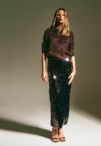 Pullover marrone peloso e gonna nera in paillettes con paillettes circolari lucide. La modella indossa sandali con tacco marroni. Sfondo neutro.