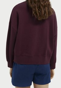 Sudadera burdeos de corte relajado, con manga larga y puños acanalados, combinada con pantalones cortos azul marino. Tejido suave y texturizado.