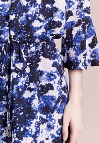 Abito con motivo blu e bianco, con vita regolabile tramite cordoncino, maniche a campana e una texture morbida. Il design presenta forme astratte.
