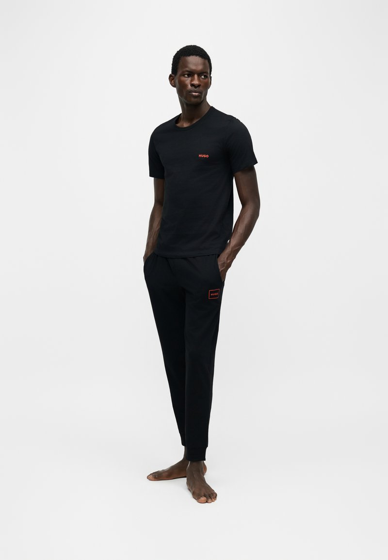 Mand stående barfodet iført sort t-shirt og joggingbukser med små røde Hugo-logoer, hænderne i lommerne, på ensfarvet hvid baggrund.