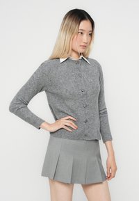 Grå strikket cardigan med lange ermer, knappet front og rund krage. Båret over en lys grå plisséd miniskjørt.