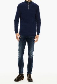 Navyn blå zip-tröja med ribbad textur, kombinerad med bleka blå jeans och bruna snörboots. Har en flerfärgad randig krage.