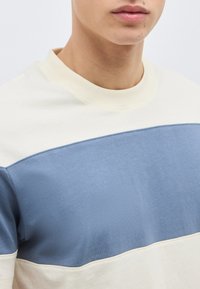 Κοντινό πλάνο ενός ατόμου που φοράει φούτερ με λαιμόκοψη χρώματος κρεμ και μπλε, εμφανίζοντας τον λαιμό, τα χείλη και το πάνω μέρος του κορμού.