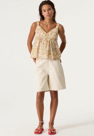 Top in cotone giallo chiaro con motivo floreale, scollo a V e spalline larghe; abbinato a pantaloncini beige fino al ginocchio e infradito rosa.