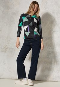 Schwarzes Oberteil mit geometrischen Mustern in Grün, Weiß und Lila; kombiniert mit navyblauen, weit geschnittenen Jeans und weißen Sneakers. Glatter Stoff, lässige Passform.