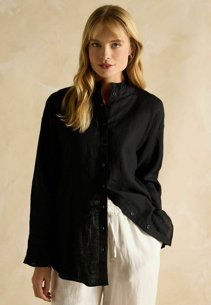 Woman with long blonde hair wearing a black button-up blouse and white drawstring pants against a neutral background.