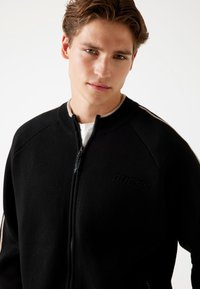 Schwarze Zip-Jacke aus strukturiertem Stoff, mit geripptem Kragen und seitlichen Streifen. Verfügt über ein dezentes, prägiertes Logo auf der Vorderseite.