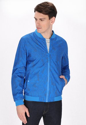 Blauwe bomberjas met een geometrisch patroon in donkerblauwe tinten, met ribbels aan de kraag, mouwen en tailleband. Model draagt het met de handen in de zakken.