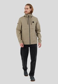 Beige wasserdichte Jacke mit einem Frontreißverschluss, verstellbarem Kapuzenaufsatz und zwei Taschen; kombiniert mit dunklen Jeans und grauem Schuhwerk.