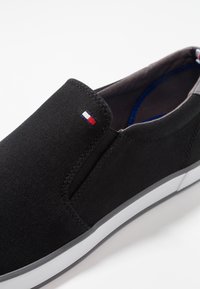 Czarne płócienne buty typu slip-on z białą gumową podeszwą, z małym akcentem trójkolorowej flagi w pobliżu pięty oraz gładką teksturą.