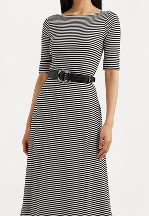 Femme portant une robe noire et blanche à rayures horizontales avec des manches jusqu'au coude, cintrée à la taille par une ceinture noire avec une boucle à double anneau.