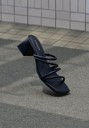 Fekete szandál négyzet alakú orral, három vékony pánttal és blokk sarkú, járdán, csempézett fal mellett.