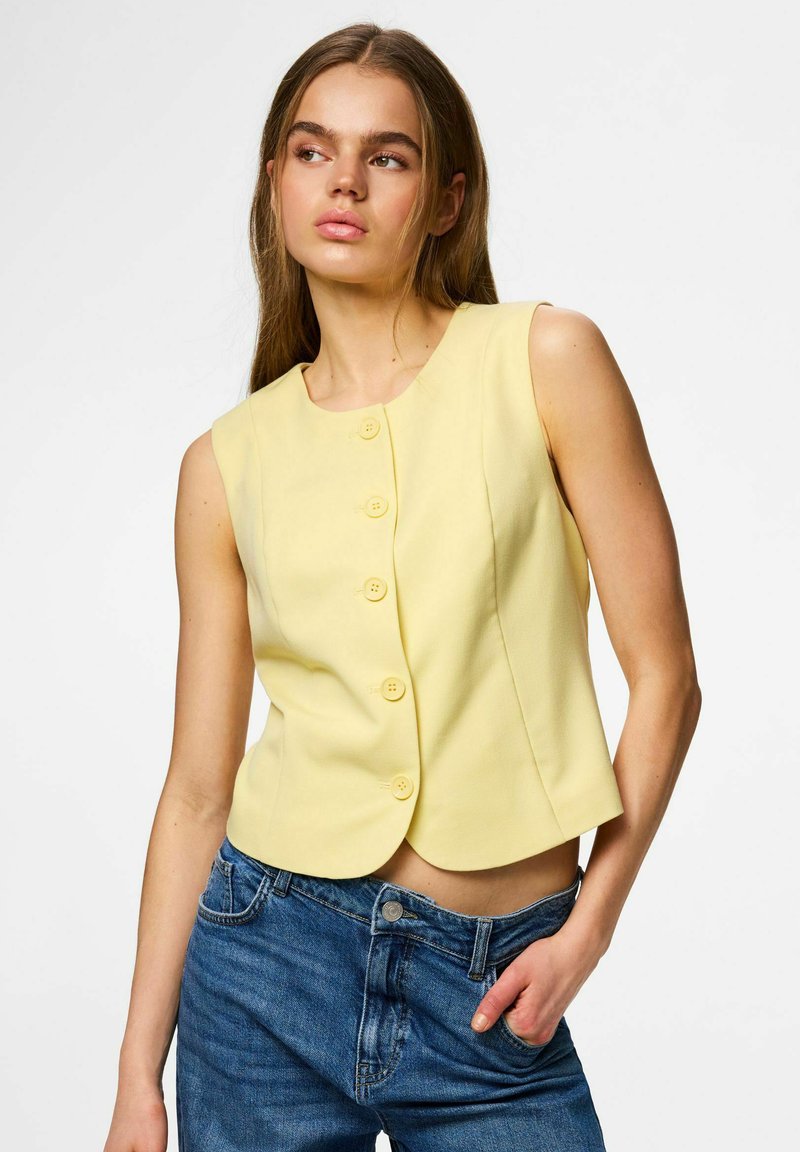 Jeune femme portant un chemisier jaune sans manches boutonné et un jean bleu, posant avec une main dans la poche, regardant sur le côté.