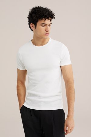 Jeune homme aux cheveux bouclés portant un t-shirt blanc à manches courtes et un pantalon noir, debout les mains dans les poches, regardant vers le bas à sa gauche.