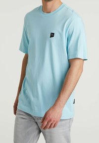 Jasnoniebieska bawełniana koszulka z krótkim rękawem i małą czarną naszywką z logo na piersi. Miękka tekstura, luźny krój i okrągły dekolt.
