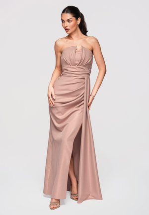 Frau posiert in einem trägerlosen, roséfarbenen Satinabendkleid mit drapiertem Oberteil, hohem Schlitz und silbernen Riemchen-High Heels vor einfachem Hintergrund.