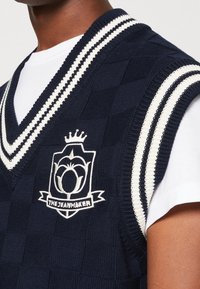 Gilet en tricot bleu marine avec un col en V et des accents à rayures blanches ; présente un logo avec un motif floral et le texte "THE JEANMAKER."