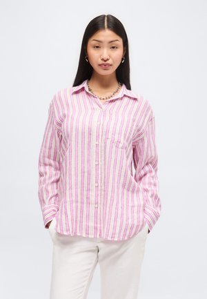 Mladá žena s dlhými čiernymi vlasmi, oblečená v ružovo-bielej vertikálne pruhovanej košeli na gombíky a bielych nohaviciach, stojaci s rukami v vreckách.