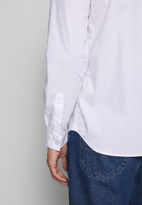 Camisa blanca de algodón con mangas largas, puños abotonados y un corte relajado, combinada con jeans de mezclilla azul. Diseño minimalista sin estampados.