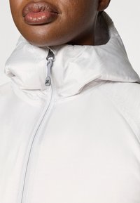 Valkoinen, kevyt takki, jossa on korkea kaulus, etuvetoketju ja hillitty brändäys. Ominaisuuksiin kuuluu sileä, kiiltävä pinta ja minimimäärä saumoja.