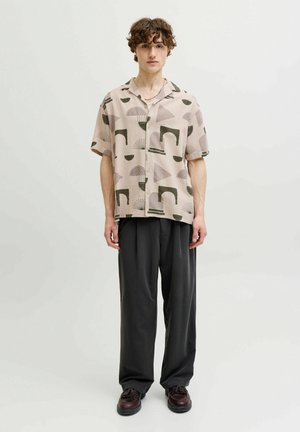 Junger Mann mit lockigem Haar, der ein beiges kurzärmeliges, gemustertes Hemd mit Knöpfen, schwarze lockere Hosen und dunkelbraune Schuhe trägt und vor weißem Hintergrund steht.