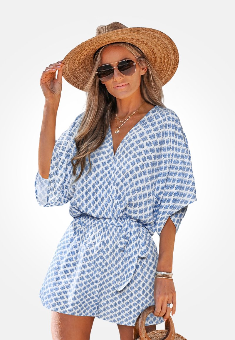 Vrouw draagt een blauw-wit motief jumpsuit, grote strohoed, zonnebril en houdt een ronde strozak met houten handvat vast.