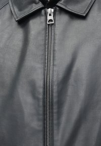 Primo piano di una giacca di pelle nera con la parte anteriore che mostra una cerniera in metallo argentato e il colletto, con texture e cuciture visibili.