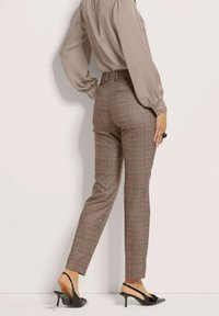 Braun karierte Hose aus einem strukturierten Stoff mit schmalem Schnitt, kombiniert mit einer hellen Bluse und schwarzen, hohen Sandalen.