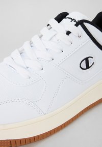 Vit sneaker med perforerad textur, svarta accenter och vita snören. Har en gummisula och en logotyp på sidan.