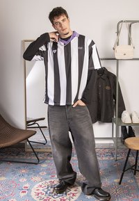 Tricou cu dungi alb-negru și guler violet, asortat cu blugi gri lejeri și pantofi negri. Ambientul camerei are tonuri neutre.