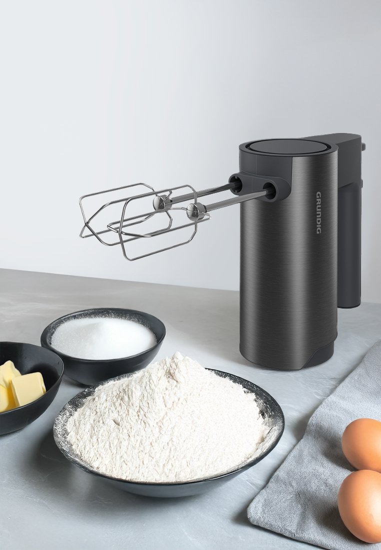 Elektrischer Handmixer in gebürstetem Dunkelgrau, mit Rührbesen aus Edelstahl. Umgeben von Schüsseln mit Mehl, Zucker, Butter und Eiern auf einer hellen Oberfläche.