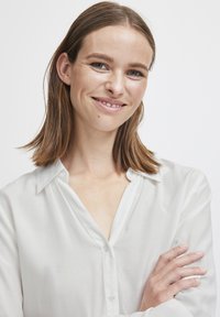 Camicia bianca leggera con colletto, caratterizzata da un leggero motivo a trama. Maniche lunghe con polsini a bottone.