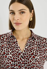 Blusa de tela ligera con base blanca y manchas de leopardo en negro y rojo, cuello abotonado y botones negros.