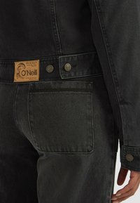 Sort denimjakke med et brunt lædermærke, metalknapper og detaljer med sydde lommer. Stofoverfladen har en struktureret finish.