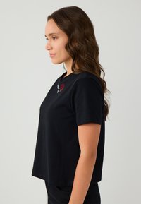 Een vrouw met lang golvend bruin haar staat zijwaarts en draagt een zwart t-shirt met korte mouwen met een klein rood en wit hart ontwerp bij de halslijn.