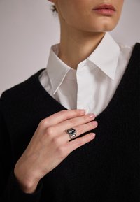 Sølvmetallring med et skulpturelt design, båret på en hånd. Bakgrunnen inkluderer en svart genser og en hvit skjorte med krage.