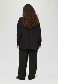 Zwarte longsleeve met een losse pasvorm en knoopmanchetten, gecombineerd met wijde zwarte broeken. Kruidige haren vallen over de schouders.