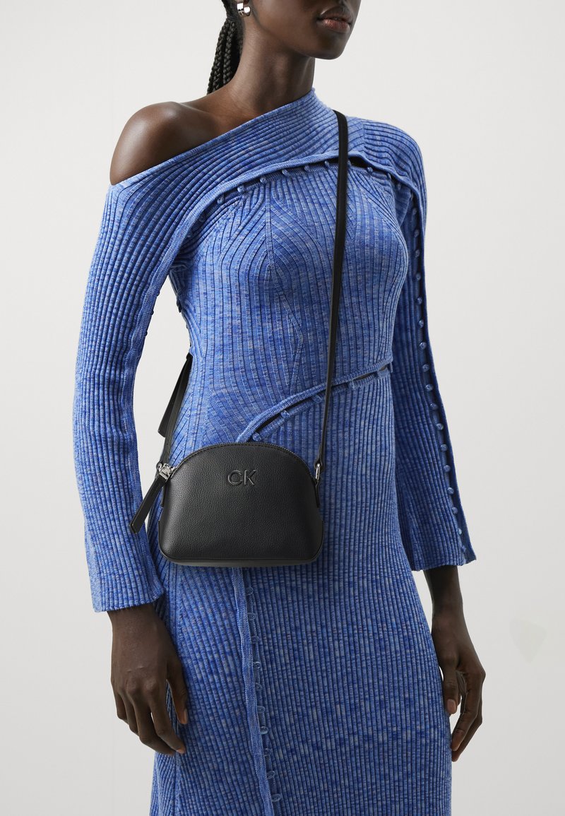 Abito a maglia blu a coste con design off-shoulder, caratterizzato da dettagli con bottoni. Piccola borsa a tracolla nera in pelle con logo impresso.
