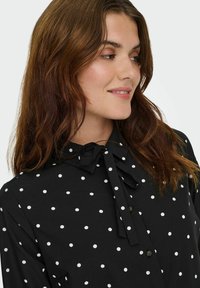 Juoda palaidinė su baltais taškeliais, priekyje su sagomis, kaklaraištis įmazgas prie apykaklės, ilgos rankovės su rankogalių detalėmis, lygi audinio tekstūra.