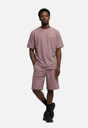 Homme portant une chemise mauve à manches courtes et un short assorti avec des boutons orange, des chaussettes noires et des chaussures noires, debout avec une main dans la poche.