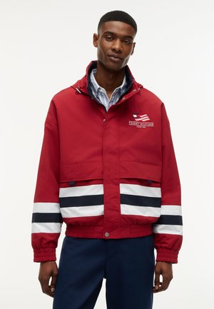 Mand iført en rød Tommy Hilfiger-jakke med hvide og marineblå striber på ærmerne og lommerne, kombineret med marineblå bukser, står foran en hvid baggrund.
