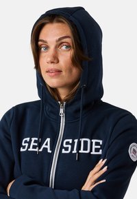 Veste à capuche zippée marine avec le lettrage blanc "SEASIDE", des cordons de serrage et un patch rond sur la manche gauche. Matériau en mélange de coton doux.