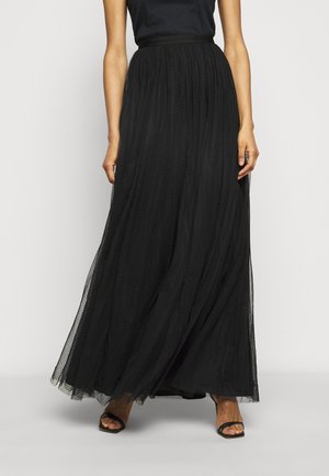 Vrouw draagt een zwarte, hooggetailleerde, tot op de grond vallende tule rok en zwarte sandaaltjes met bandjes, tegen een effen achtergrond.