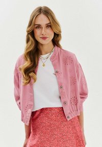 Różowa bombowiec z haftem kwiatowym, wykończona srebrnymi guzikami; nałożona na biały top i wzorzystą różową spódnicę.