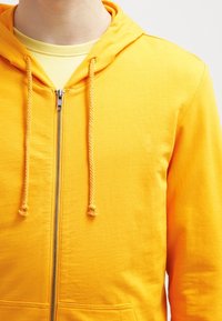 Sudadera amarilla con capucha, cremallera y cordones, con una textura suave, que presenta un bolsillo frontal y está combinada con una camiseta de color amarillo claro debajo.