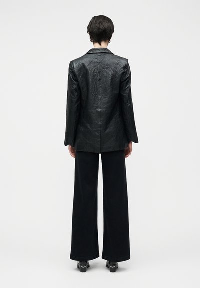 Μαύρο δερμάτινο σακάκι με υφή, με μία σκίση στην πλάτη και μακρύ μανίκι, συνδυασμένο με μαύρο παντελόνι φαρδιάς γραμμής και παπούτσια με τακούνι.