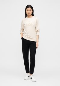 Μπεζ φούτερ με λογότυπο "EA7", συνδυασμένο με μαύρο παντελόνι. Το μοντέλο φοράει λευκά αθλητικά παπούτσια με μαύρες λεπτομέρειες. Ομαλό ύφασμα και άνετη εφαρμογή.