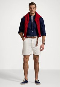 Κόκκινο γιλέκο με καπιτονέ ύφασμα πάνω από σκούρο μπλε πουκάμισο με κουμπιά, ριγέ μπλουζάκι, λευκά σορτς, καφέ πλεκτή ζώνη και σκουρομπλε παπούτσια.